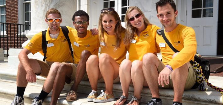 four orientation staff students wearing yellow shirts sit on steps with arms around each other