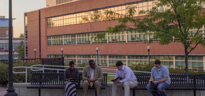 Four people sit on a bench in front of Science Hall at sunset.