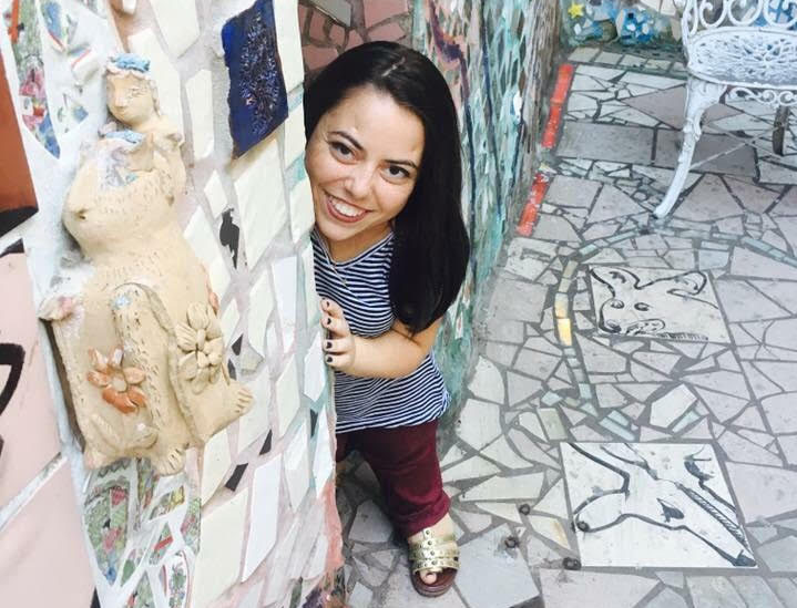 Mackenzie Trush smiles from behind a wall filled with mosaic art