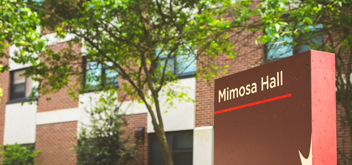 brown sign for Mimosa Hall, in front of building with blooming green trees