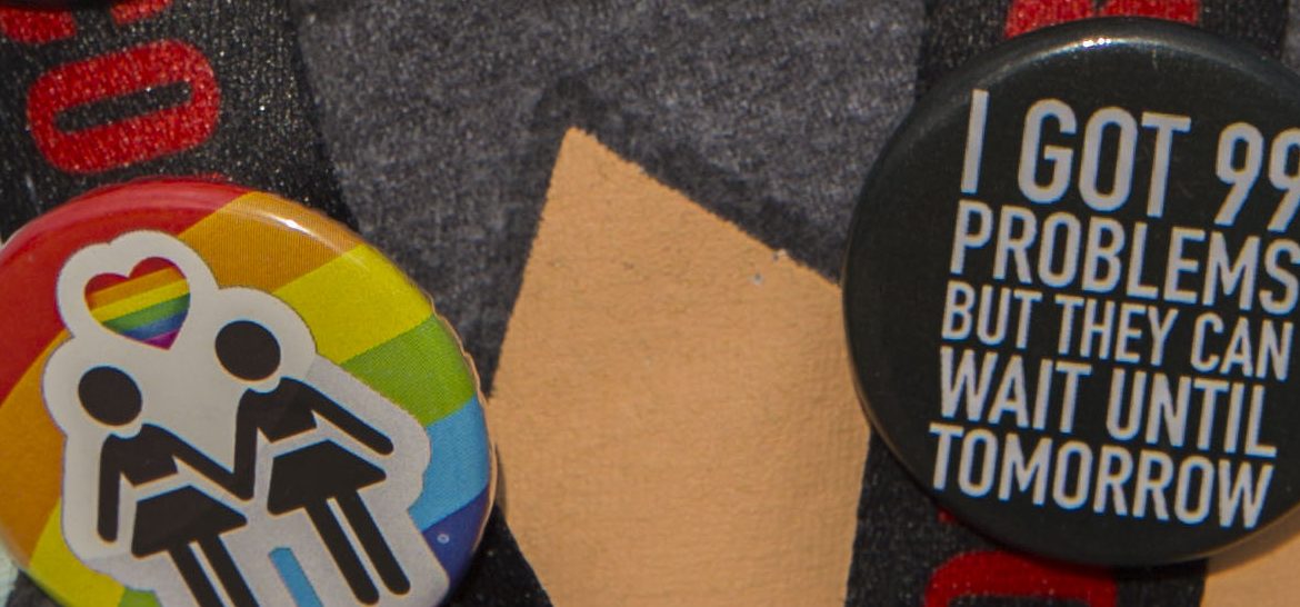 close up of buttons on students lanyard - rainbow button showing black colored stick figures in love and black button that says I got 99 problems but they can wait until tomorrow