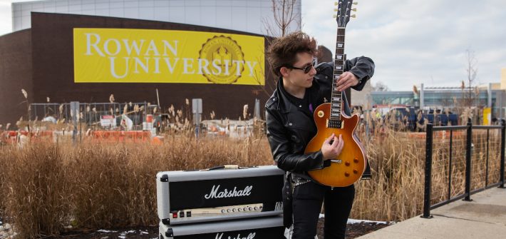 Luke outfront of Wilson Hall with his guitar and amp outfront Rowan University sign