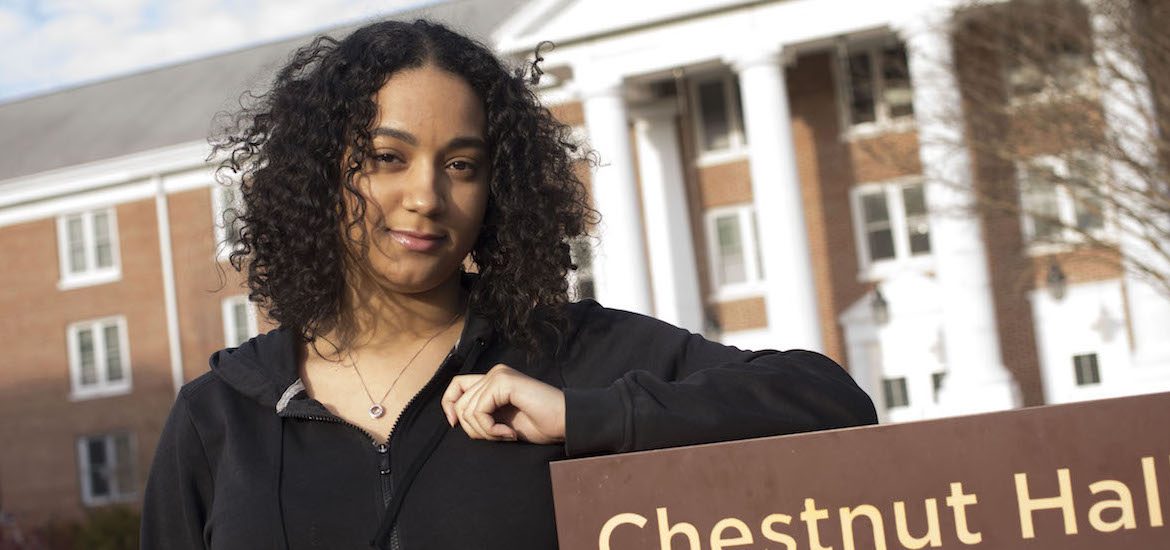 Vanessa from Rowan University stands in front of the sign for Chestnut Hall dorm, with her left elbow resting on the brown sign
