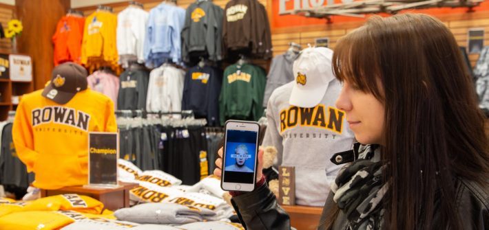 Jen Green stands in the Rowan University bookstore, looking toward her phone with the Badflower album cover OK I'm Sick