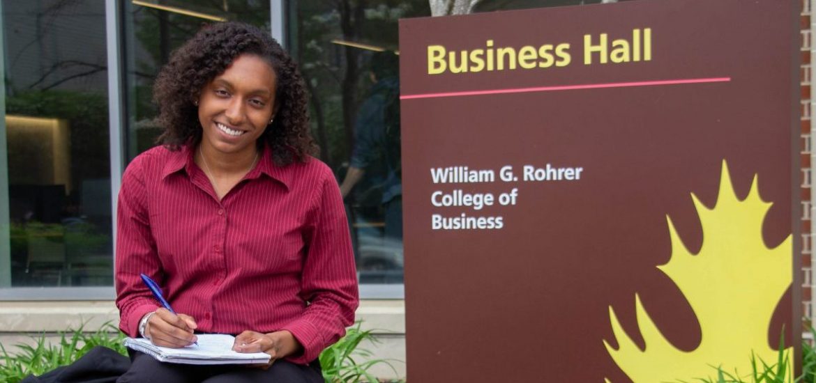 Jo Carter sits next to a Business Hall sign at Rowan University, wearing a pink button down blouse and holding a notebook