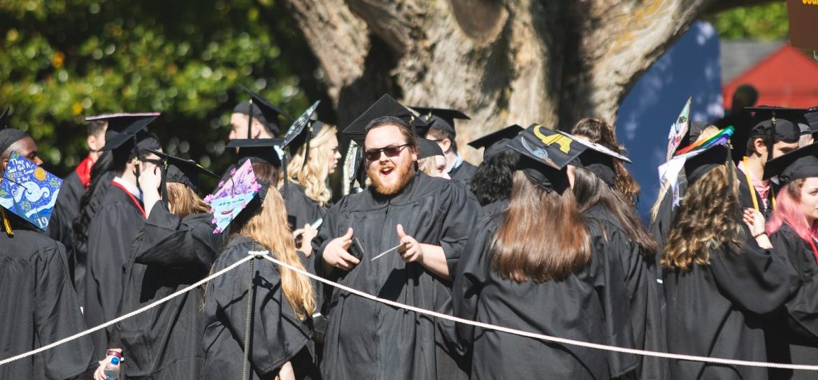 Johnathan Puglise smiles at the camera while being surrounded by other recent graduates in their black cap and gowns.
