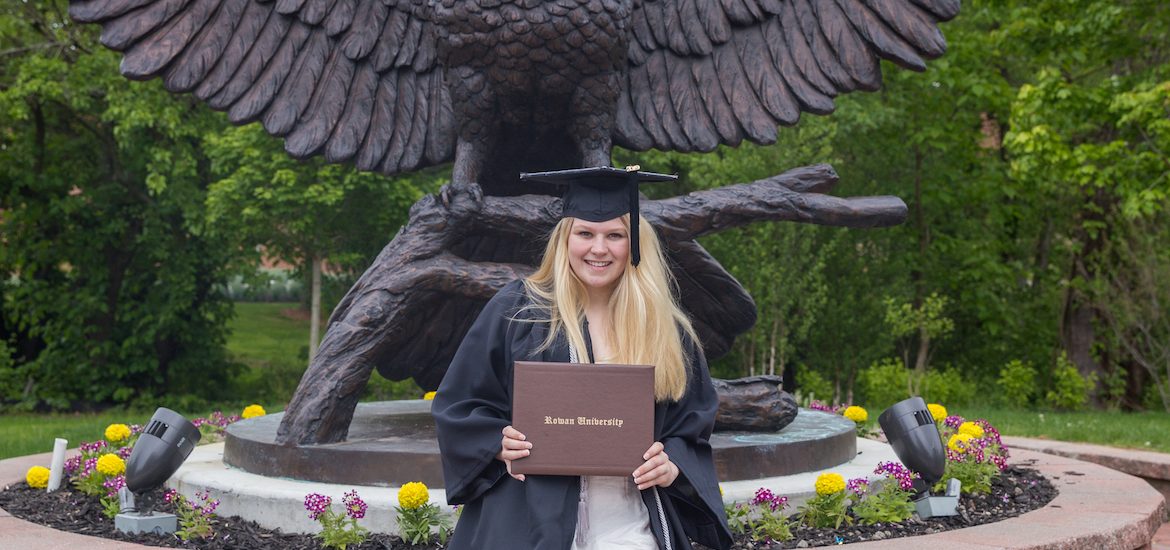 Cheyenne wears her graduation gown, posing with Rowan University diploma holder in front of owl statue