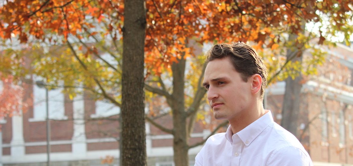 Adam looks into the distance, while wearing a crisp white button down shirt and fall colored trees behind him.