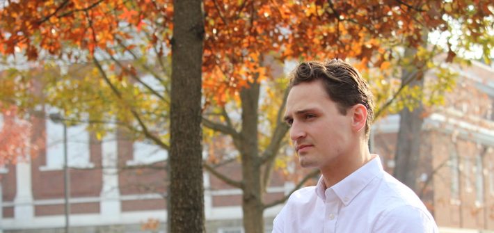 Adam looks into the distance, while wearing a crisp white button down shirt and fall colored trees behind him.