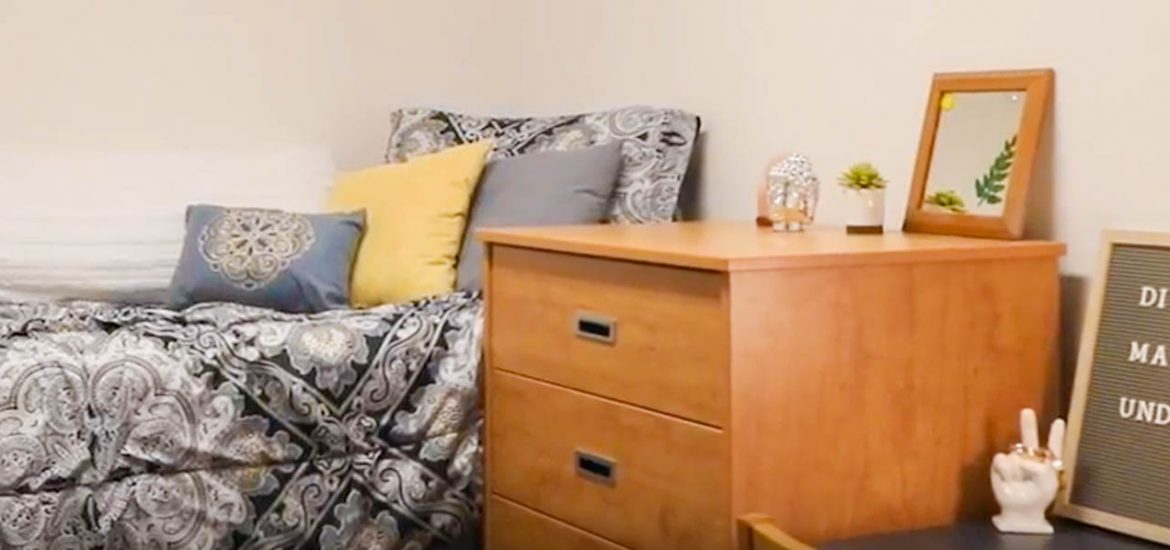 dorm room with gray comforter on bed and wooden desk and drawers.