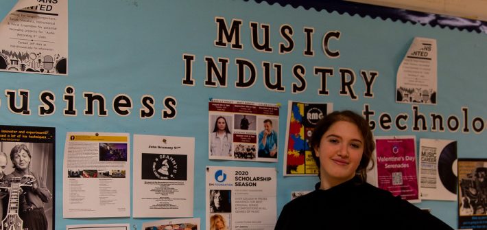 Bianca stands in front of a Music Industry bulletin board.