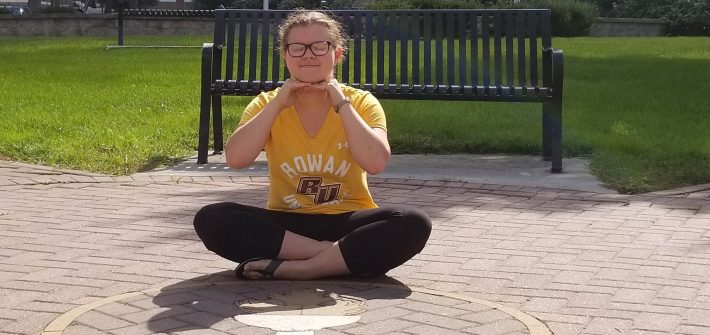 Amanda wears a Rowan shirt and sits on the ground smiling in front of a Rowan logo on the ground.