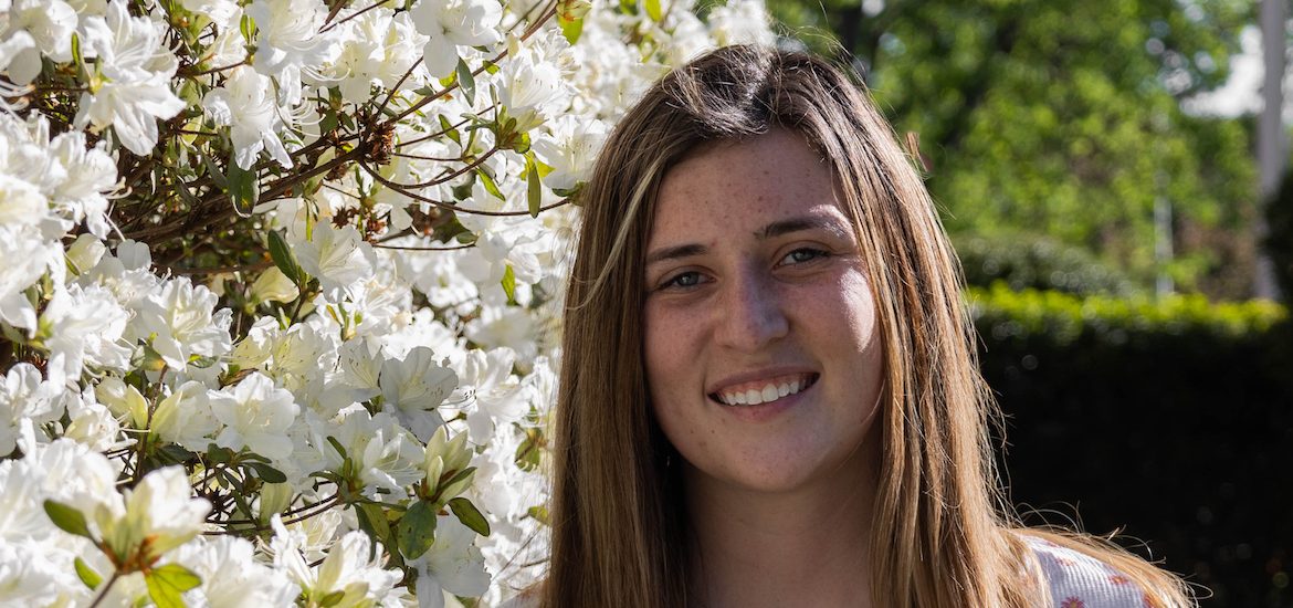 Hannah smiles while standing next to a white flowering plant on campus.