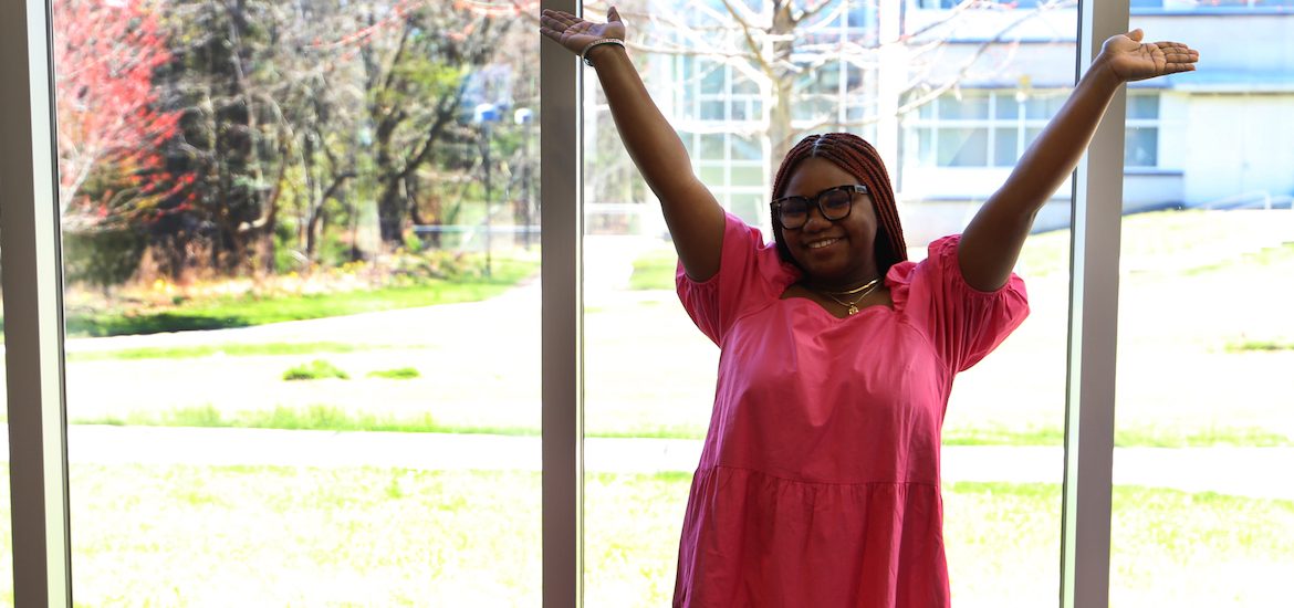 Dee poses ecstatically in a pink dress and glasses, with her hands up in the air.