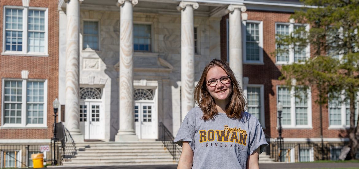 Leah smiles in front of Bunce Hall while wearing a gray Rowan shirt and glasses.