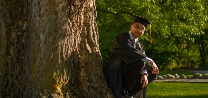 Riel wears his graduation regalia and squats by a tree.