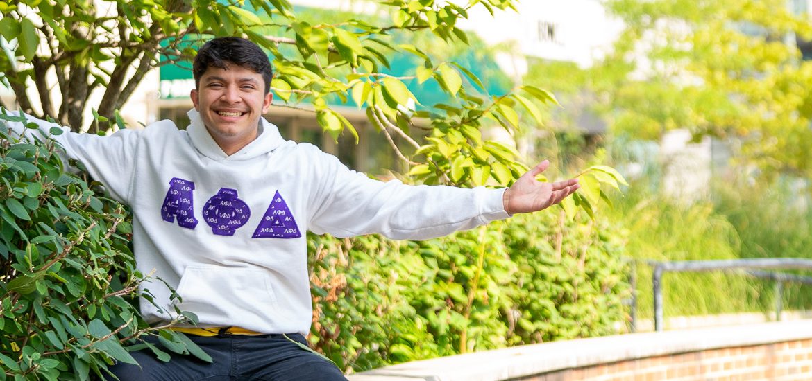 Jeremy is sporting a sweatshirt with his fraternity letters on it and is sitting down in some greenery with his arms spread open.
