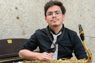 Rowan University student Jovan Rivera posing inside of Wilson Hall with a saxophone sitting in front of a piano.