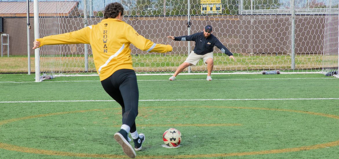 Miranda kicks a soccer ball into a soccer net as an alumnus, while wearing Rowan gold.