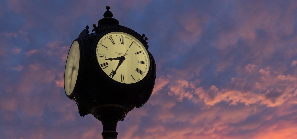 Close up of the face of a town clock with a dramatic sunset in the background.