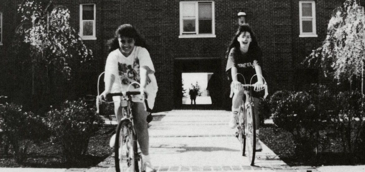 Black and white photo of two girls on bicycles outside a dorm.
